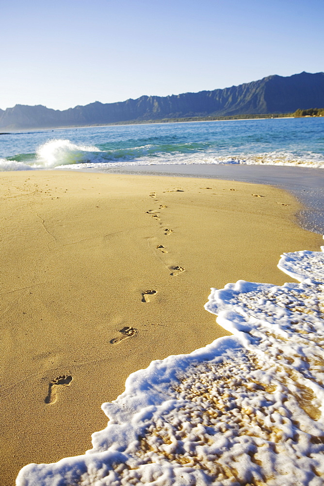 Hawaii, Oahu, Mokulua Island, footprints in the sand, water lapping on the beach, Koolau mountains in background.