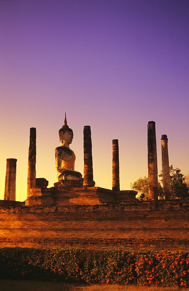 Thailand, Sukhothai, Wat Mahathat, Buddha statue with many pillars at sunset, glowing sunlight on structure.