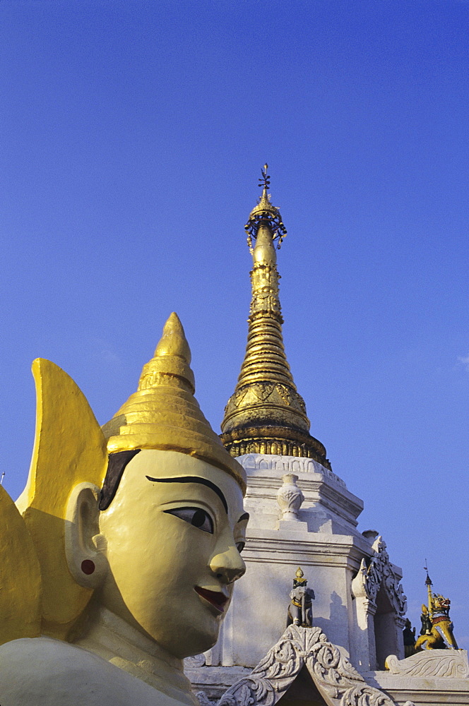 Burma (Myanmar), Yangon, Schewedagon Paya, close-up of Buddha statue and top of temple against blue sky.