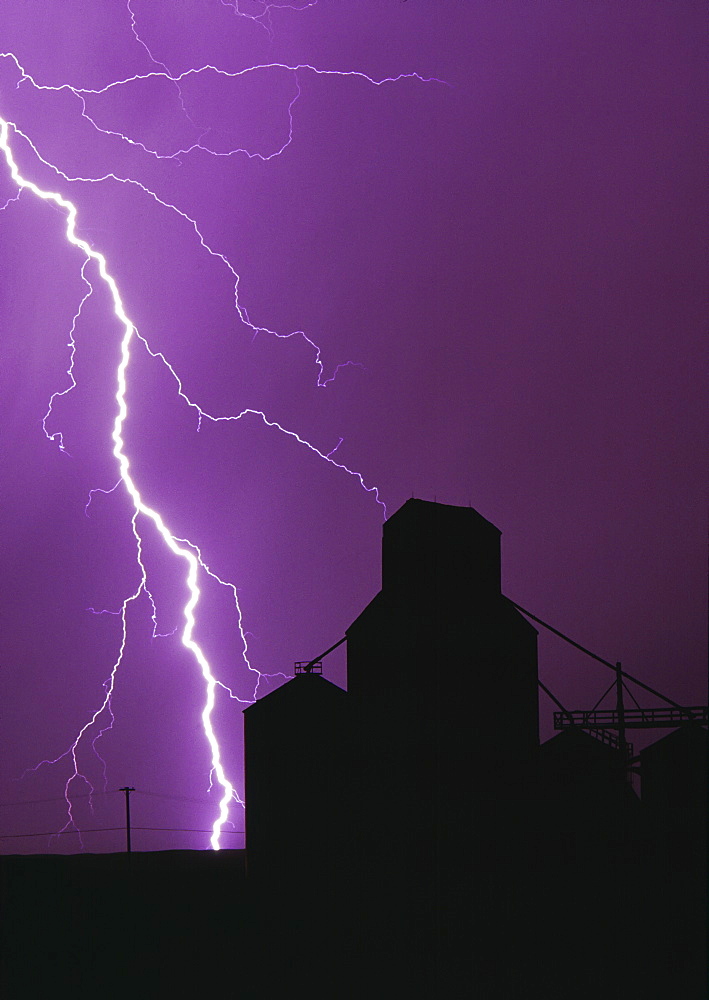 Agriculture - A bolt of lightning lights up the night sky during a storm silhouetting a grain elevator / Manitoba, Canada.