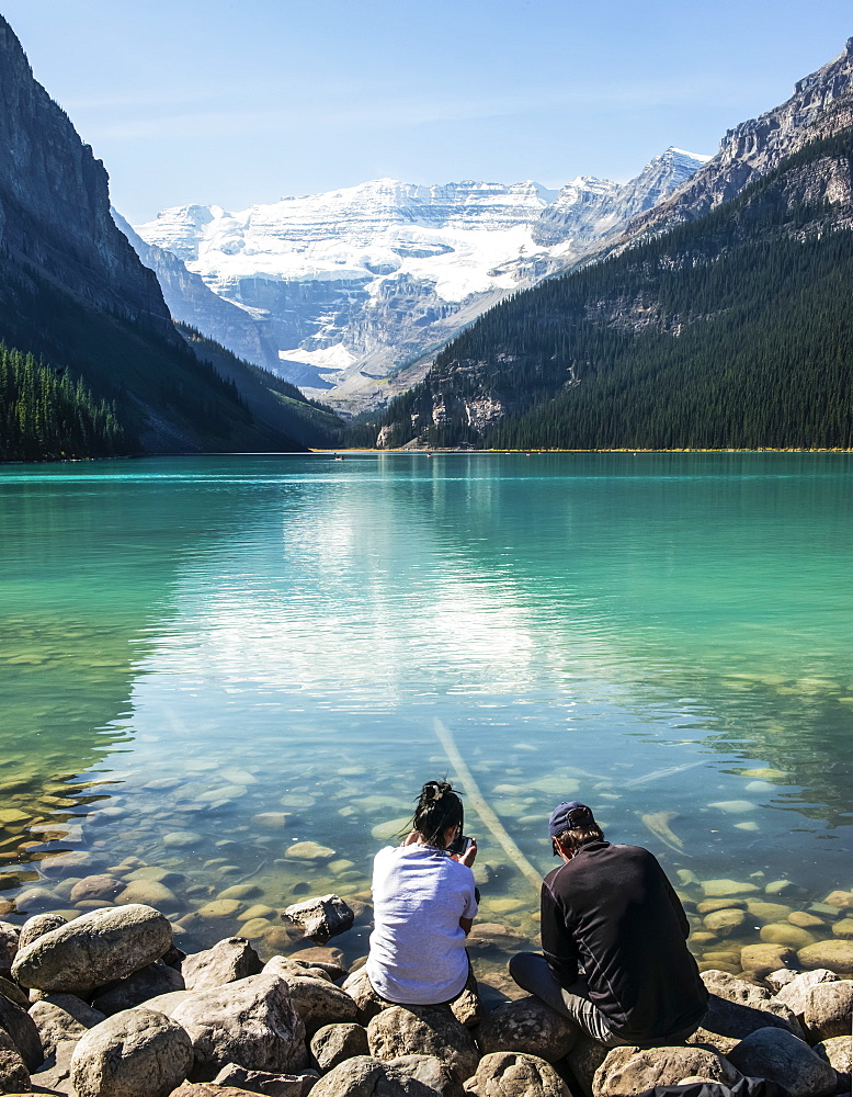 Two people distracted by looking at their smart phones while sitting along the shoreline of Lake Louise, Alberta Canada