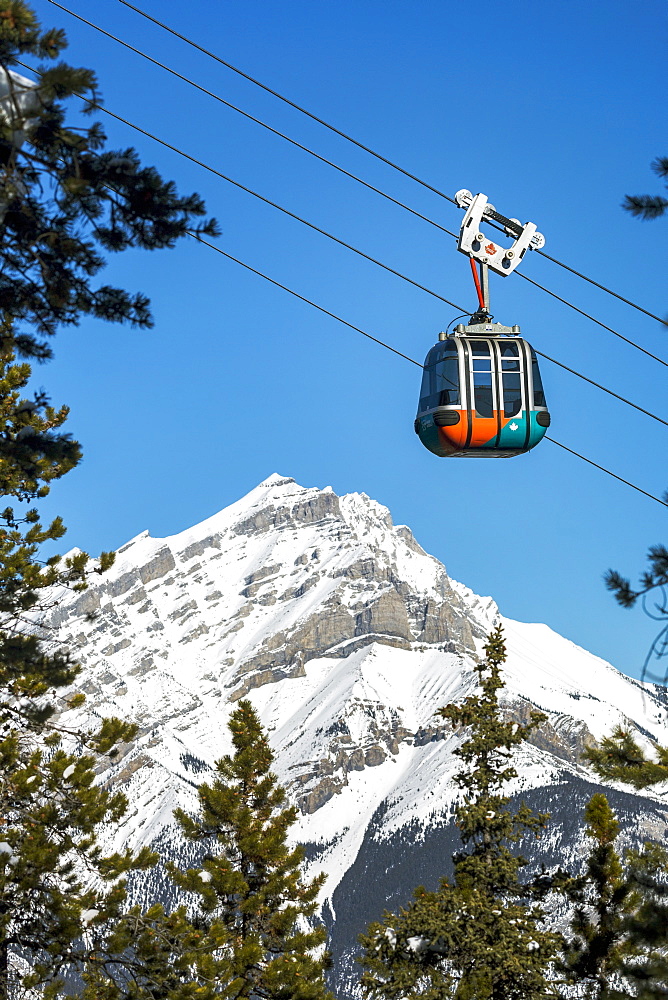 Gondola pod travelling along cables with snow covered mountain and blue sky in the background framed by evergreen trees, Banff National Park, Banff, Alberta, Canada