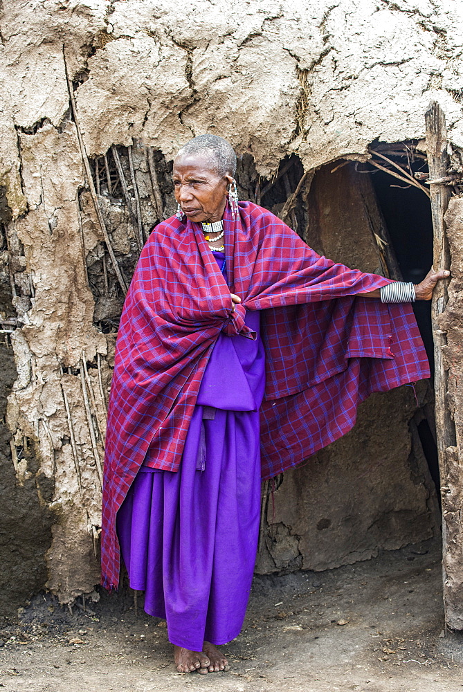 Maasai woman elder wearing traditional clothing stands at entrance to her mud hut in the Ngorongoro Conservation Area, Tanzania