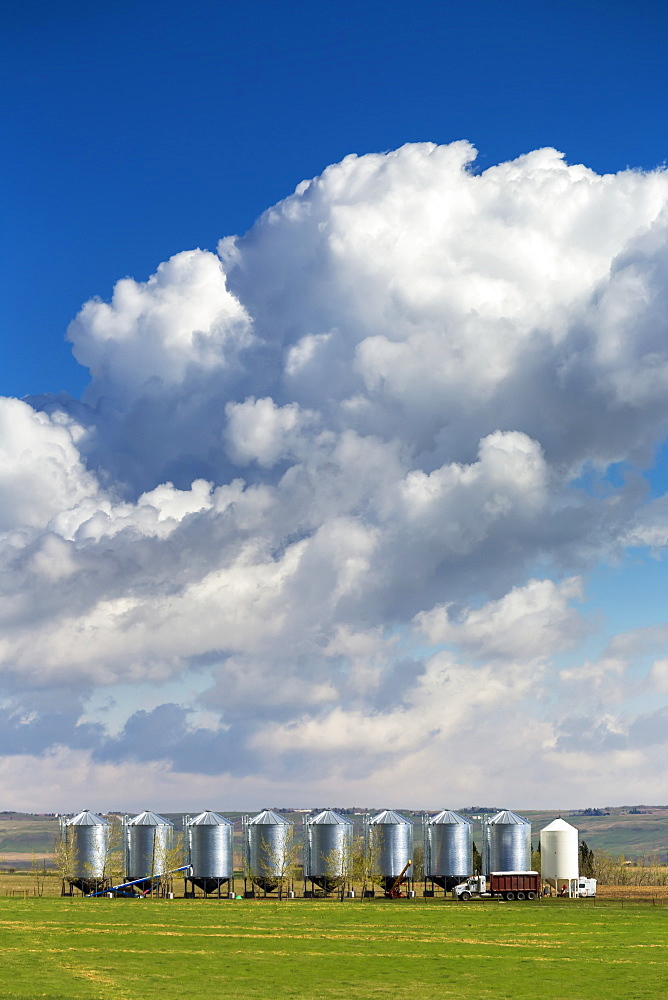 A row of large metal grain bins with dramatic storm clouds and blue sky in the background, West of Calgary, Alberta, Canada