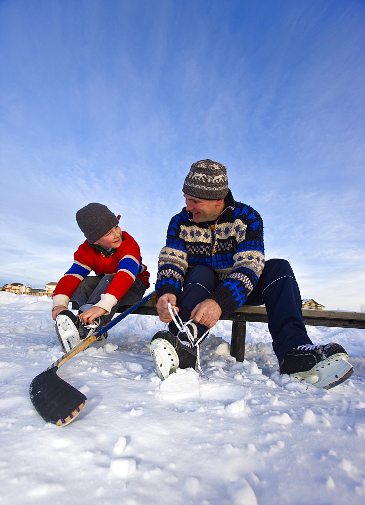 A father and son lacing their skates on a bench on a frozen lake for an outdoor hockey game, Alberta, Canada