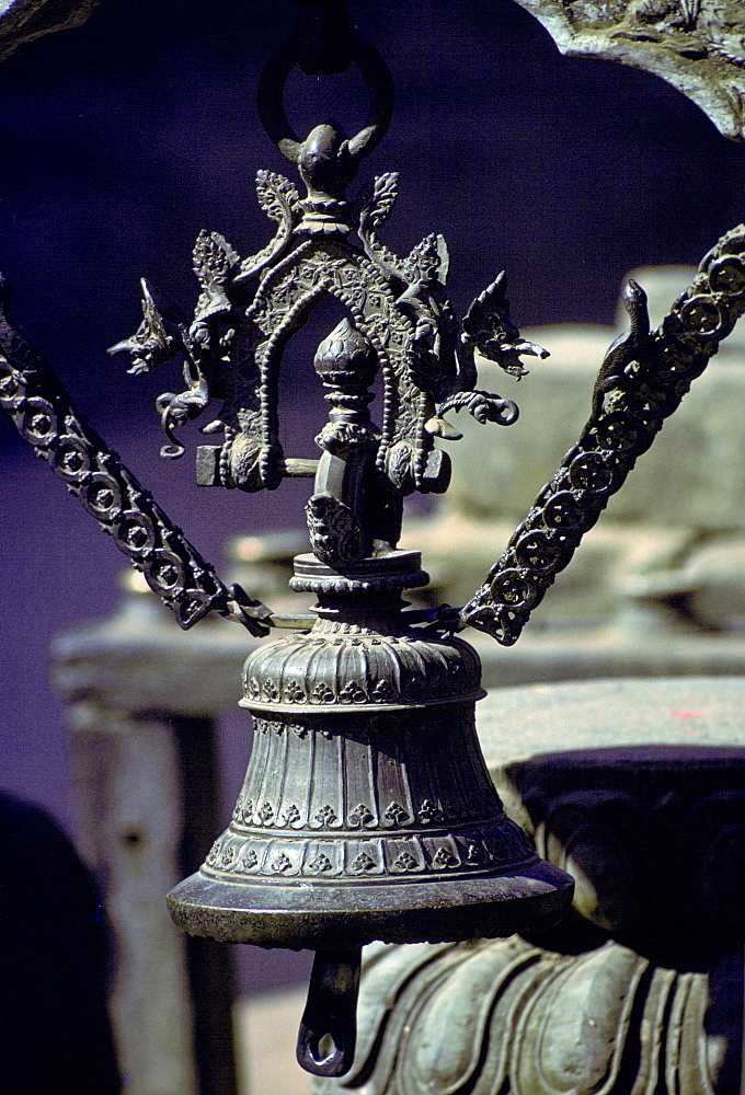 A close-up of an ornate metal bell decorated with dragon-like creatures and suspended in an arch by two metal chains decorated with lizards in Patan, Nepal.