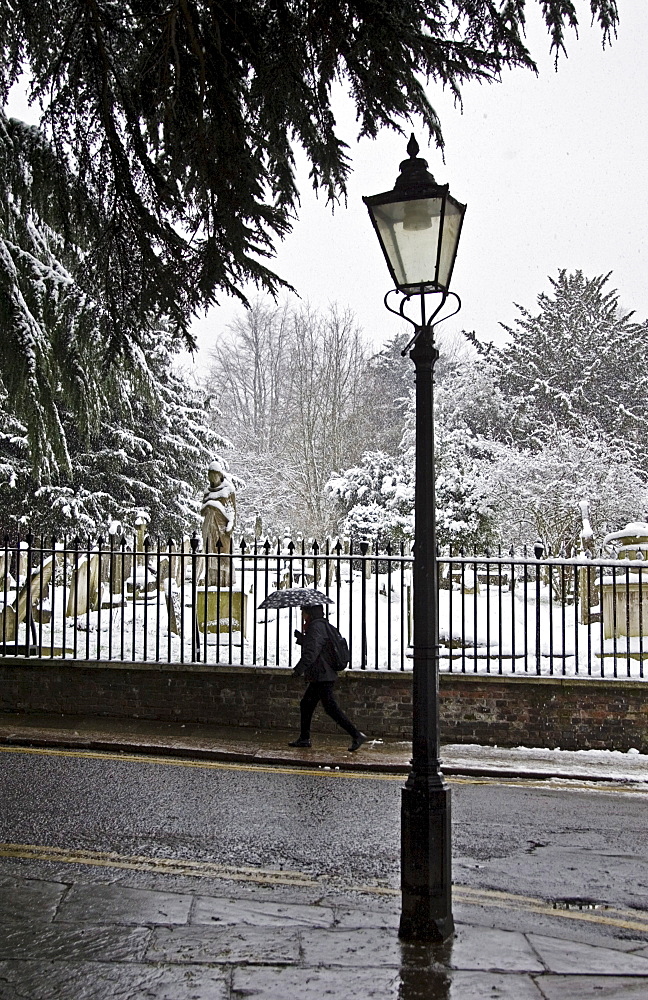 Woman with umbrella walks past snow covered graveyard, Hampstead, North London, United Kingdom