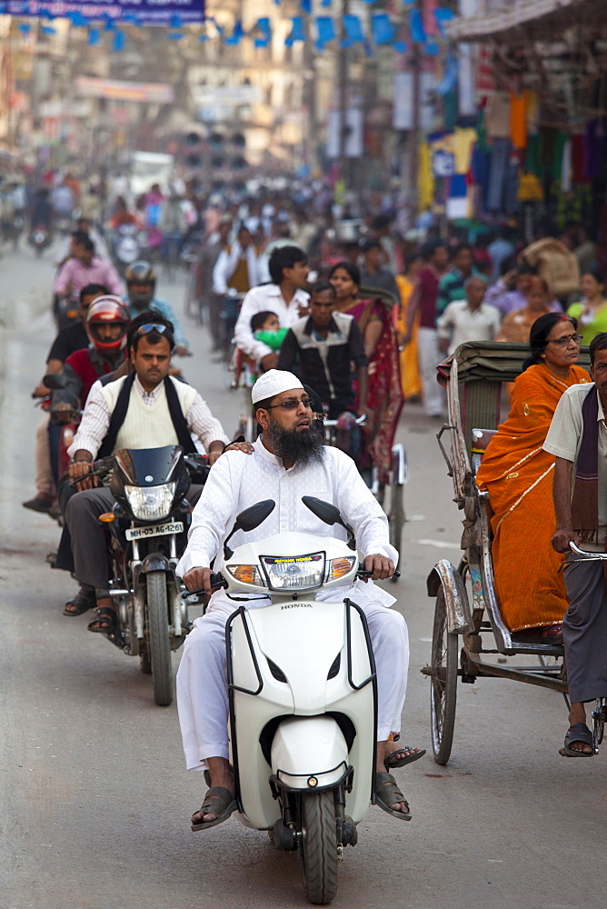 Muslim man wearing topi cap and white outfit driving motor scooter in street scene in city of Varanasi, Benares, Northern India