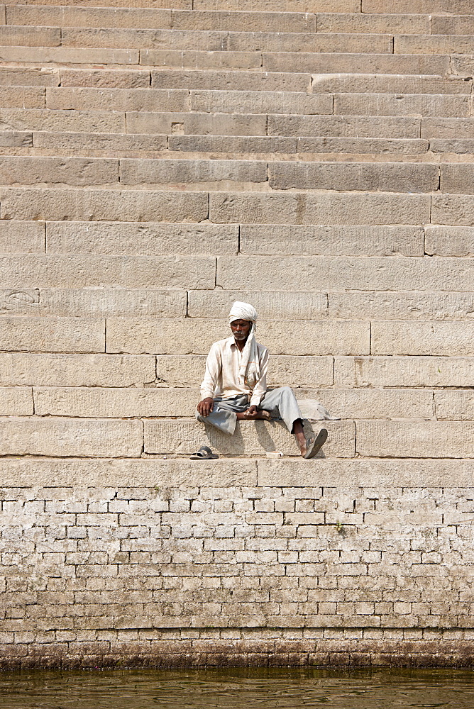 Hindu man sits on steps of Chet Singh Ghat on banks of The Ganges River in holy city of Varanasi, India