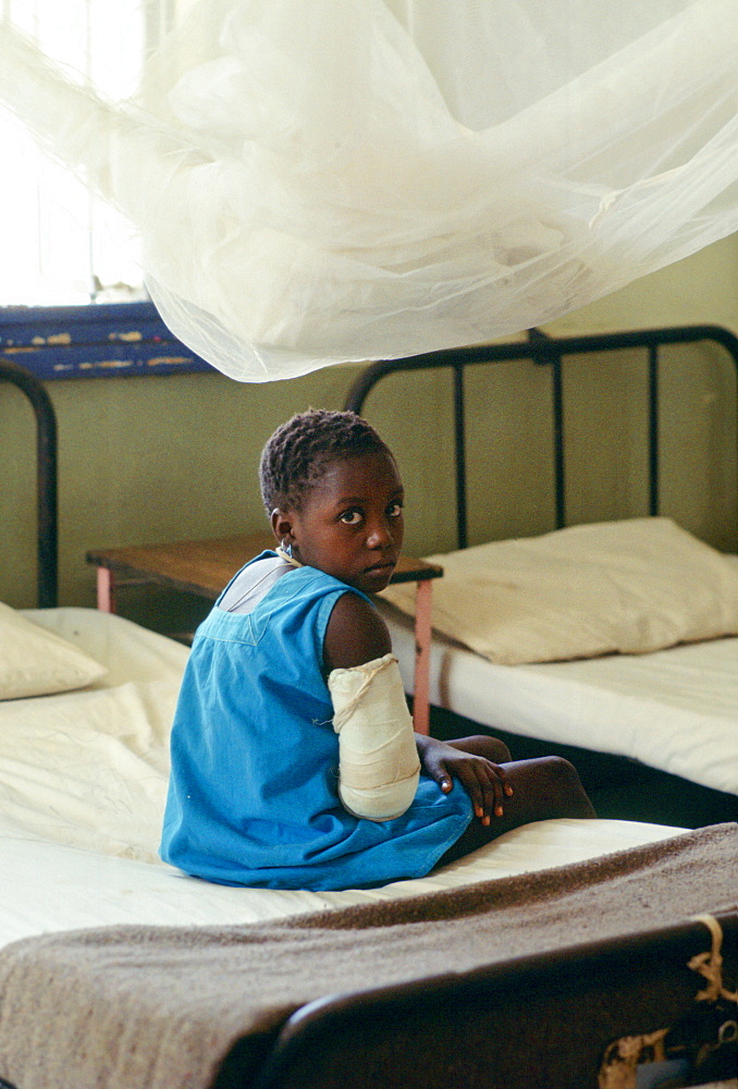 Young girl with arm in plaster bandage sits on a bed in a rural hospital supported by  the Save the Children Fund charity in The Gambia, West Africa.  A mosquito net hangs above.