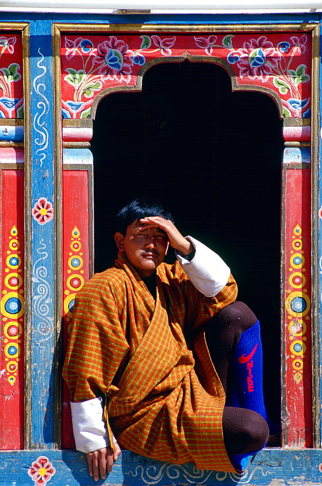 Bhutanese man shielding his eyes from the sun as he sits in an ornate carved and decorated window.  His modern Nike sports socks contrast with his traditional outfit, Paro, Bhutan