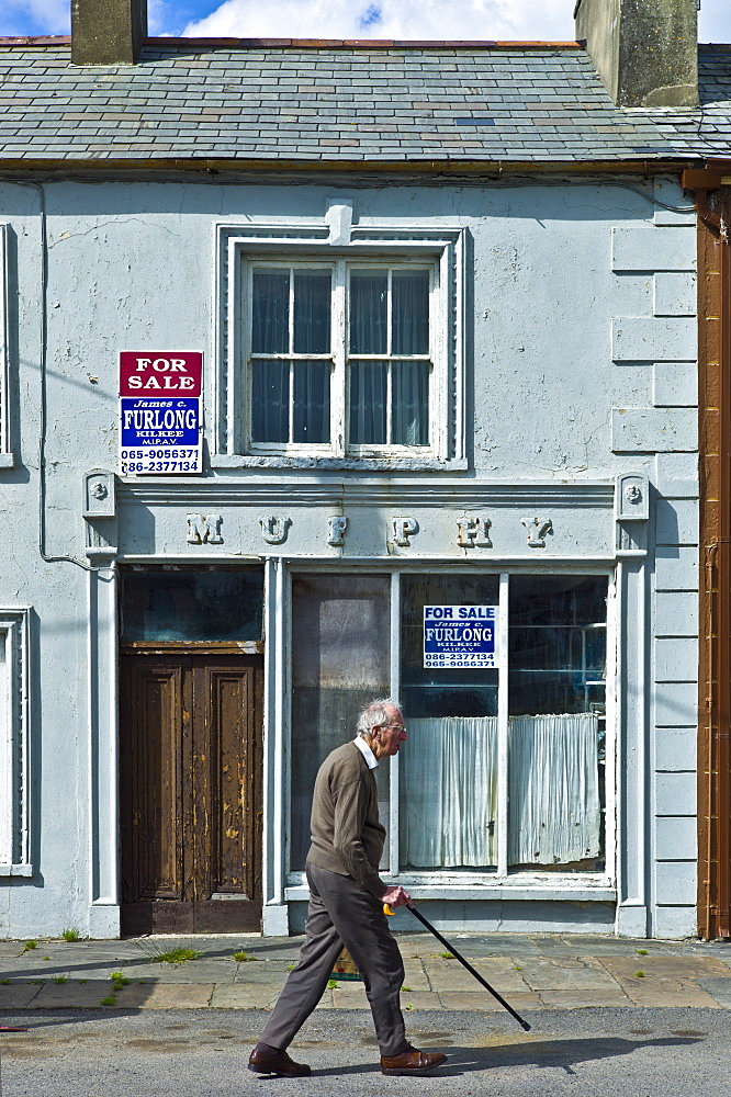 Old man strolls past Murphy shop with estate agent For Sale boards in, Kilkee, County Clare, West of Ireland