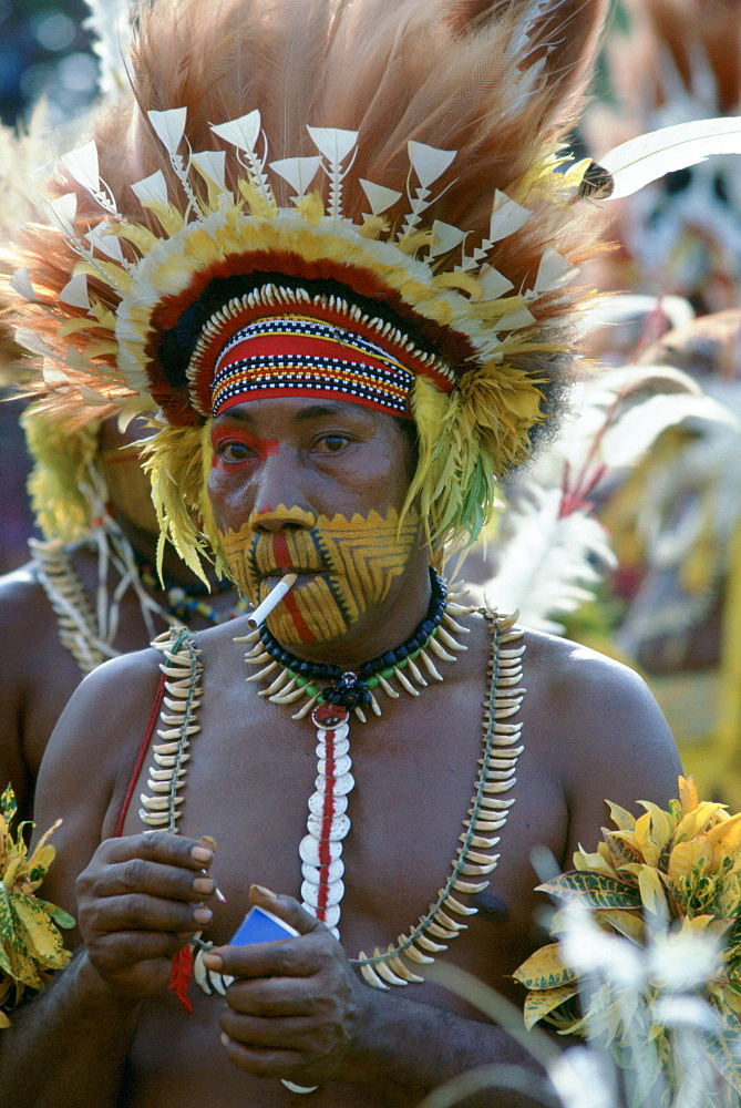 Tribesman smoking a cigarette while wearing face paints and feathered headdress during  a gathering of tribes at Mount Hagen in Papua New Guinea