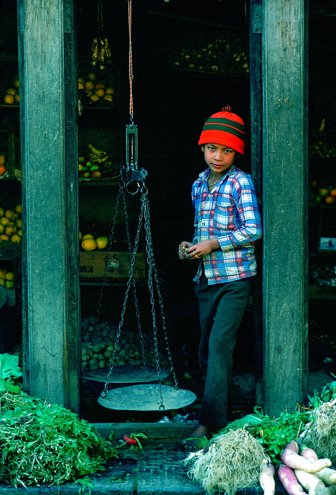 Young boy shopkeeper in a fruit and vegetables shop in Patan, Nepal.  He has old-fashioned scales for weighing the produce.