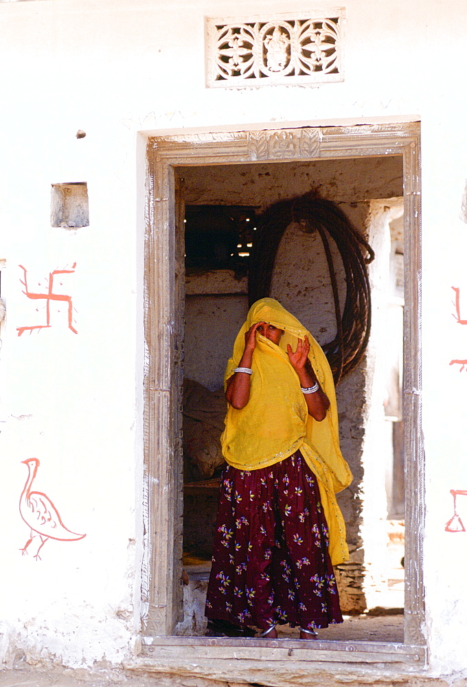 Woman villager hiding face with veil in doorway of home in Nalu, Rajasthan, India. The Swastika is a ancient symbol of good luck in India.