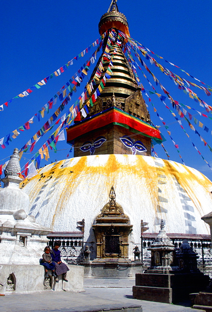 Children sitting on the wall outside theSwayambhunath Stupa  in Kathmandu valley in Nepal which is bedecked with flags.  It is a sacred monument in the Buddhist world and is a symbolic representation of the fully enlightened mind and the path to enlightenment.