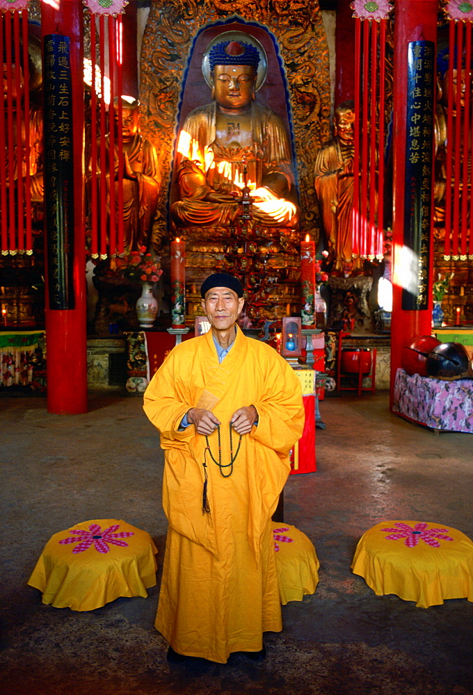 A Buddhist monk in saffron coloured robes praying in front of a statue of Buddha at the Buddhist Temple in Huating, China