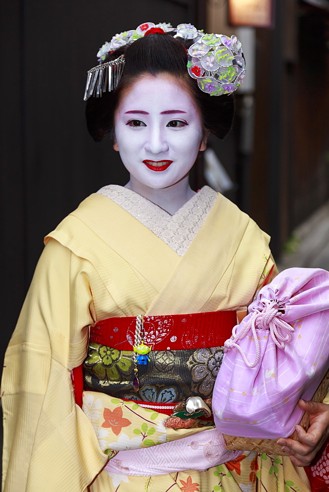 Smiling maiko, apprentice geisha, with yellow robes, stops in street on way to evening appointment, Gion, Kyoto, Japan, Asia