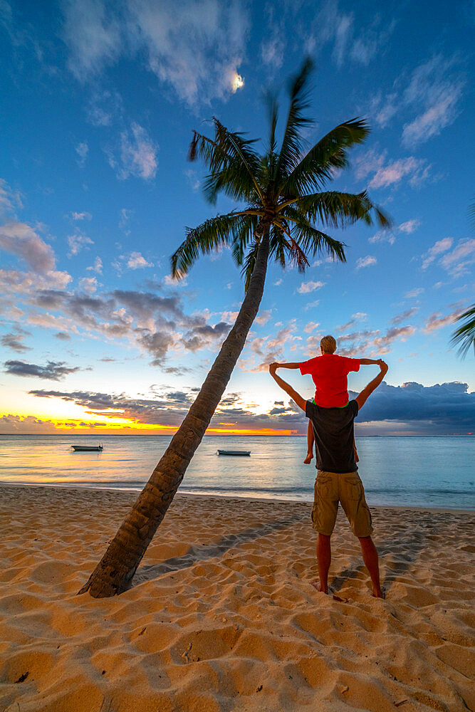 Rear view of cheerful father holding his son on neck standing on tropical sand beach, Le Morne Brabant, Mauritius, Indian Ocean, Africa