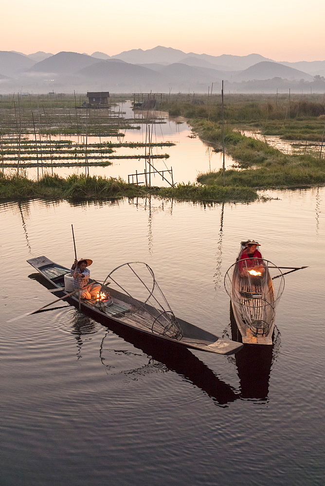 Two fishermen keep warm in their long tail fishing boats at dawn near the floating gardens on Inle Lake, Shan State. Myanmar (Burma), Asia