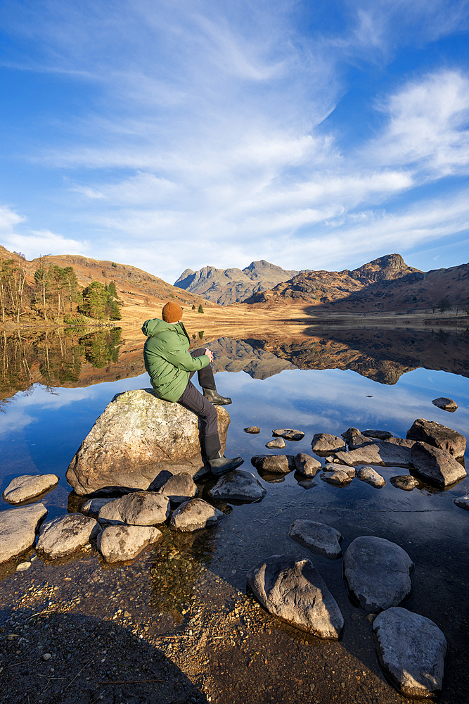Walker wearing green jacket sat admiring the mirror conditions at Blea Tarn, Lake District, Cumbria