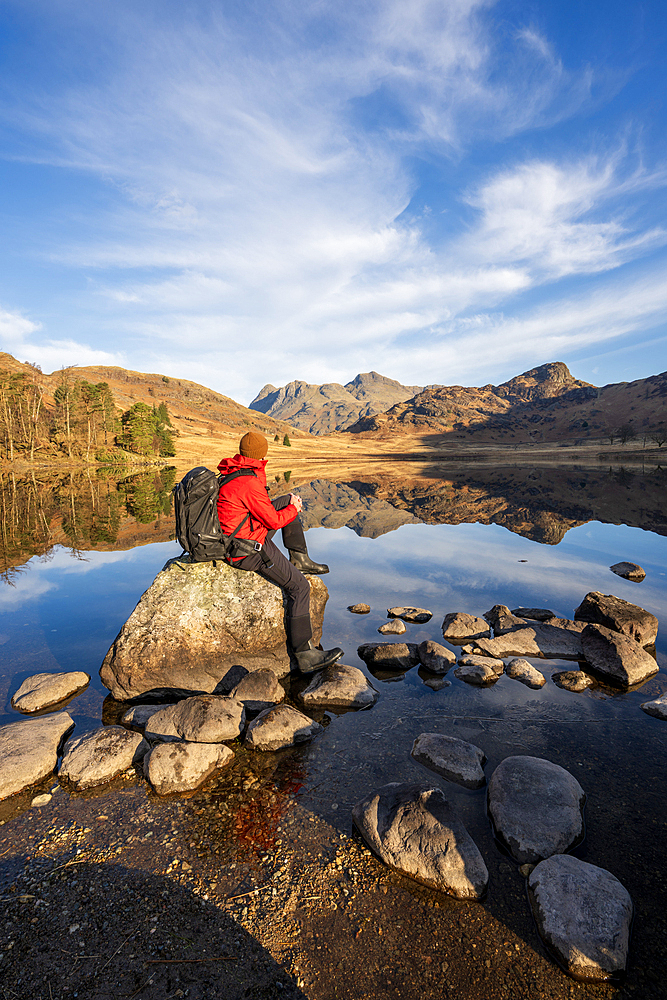Walker wearing red jacket and backpack sat admiring the mirror conditions at Blea Tarn, Lake District, Cumbria