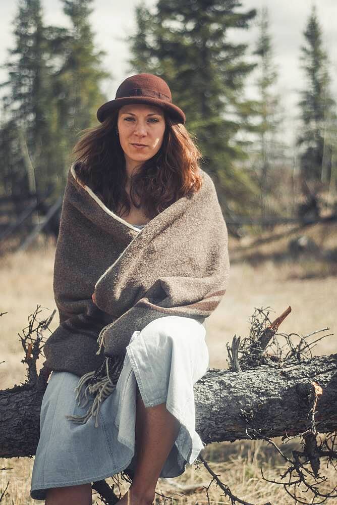 An attractive young woman sits on a log wrapped in a woollen blanket. Yukon Territory, Canada