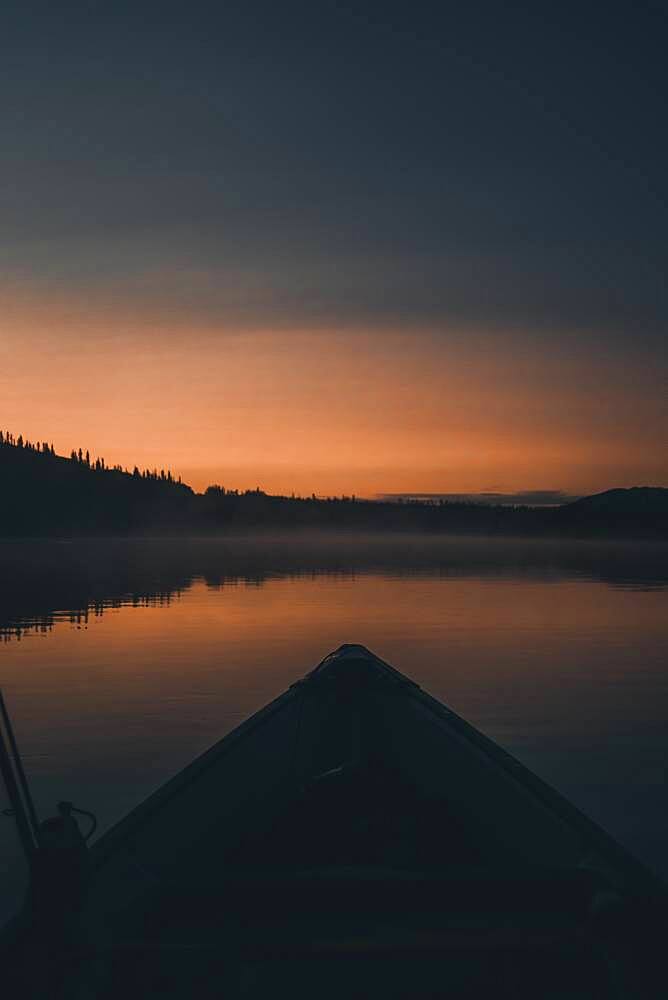 Long before the sun rises the horizon glows orange and a canoe floats over the quiet water. Yukon Territory, Canada.