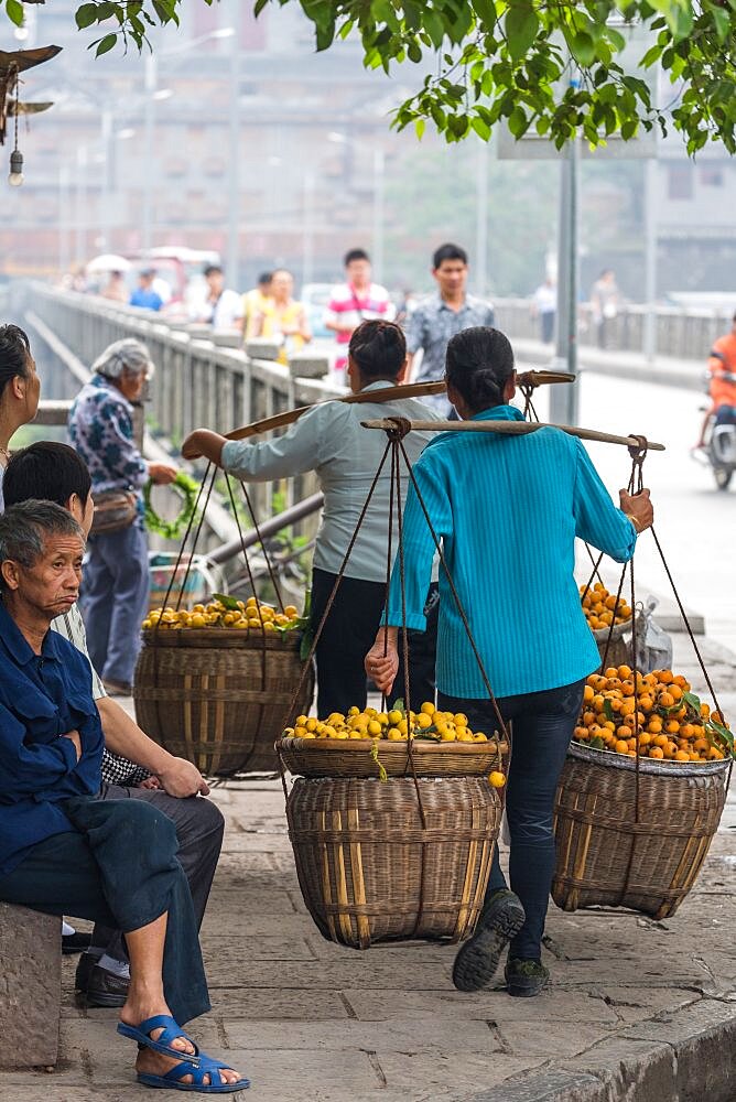 Two women carrying large baskets of fruit on sholder yokes on a street in Fenghuang, China.