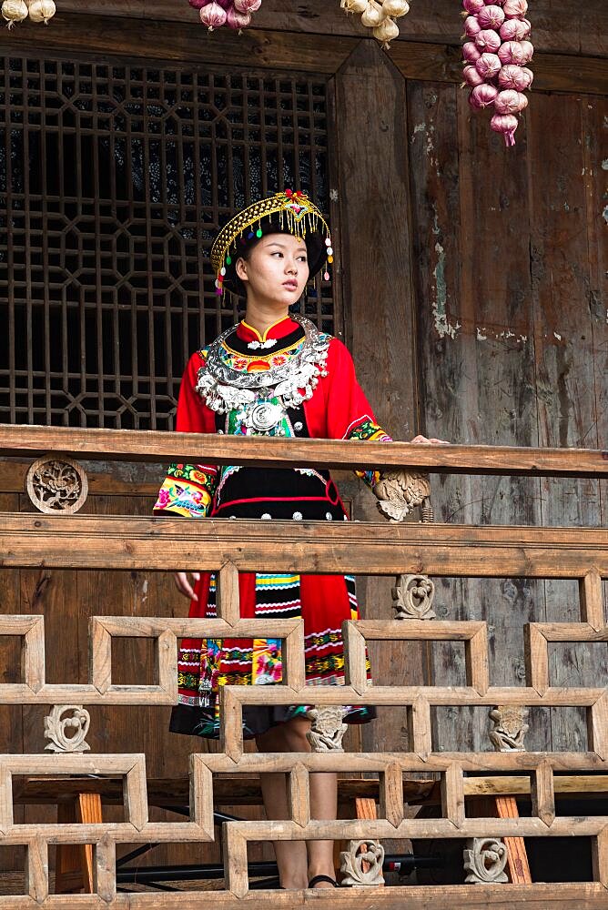 An attractive young woman in traditional ethnic Tujia dress poses for a portrait in Furong, China. Bundles of colored onions hang from the eaves as decoration.