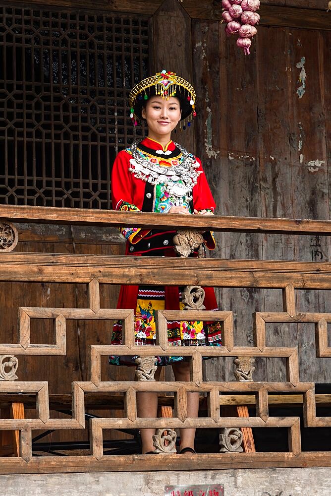 An attractive young woman in traditional ethnic Tujia dress poses for a portrait in Furong, China. Bundles of colored onions hang from the eaves as decoration.