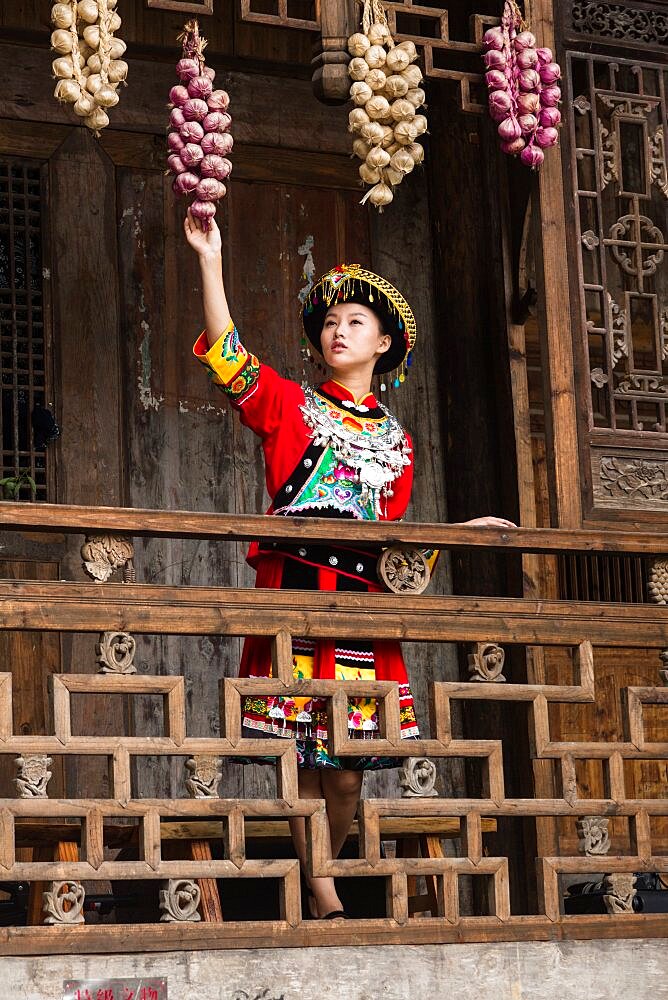 An attractive young woman in traditional ethnic Tujia dress poses for a portrait in Furong, China. Bundles of colored onions hang from the eaves as decoration.