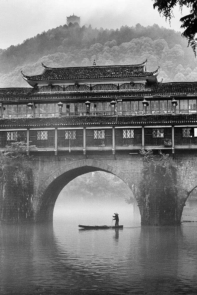 A man paddles a sampan in the fog by the Phoenix Hong Bridge on the Tuojiang River, Fenghuang, China.