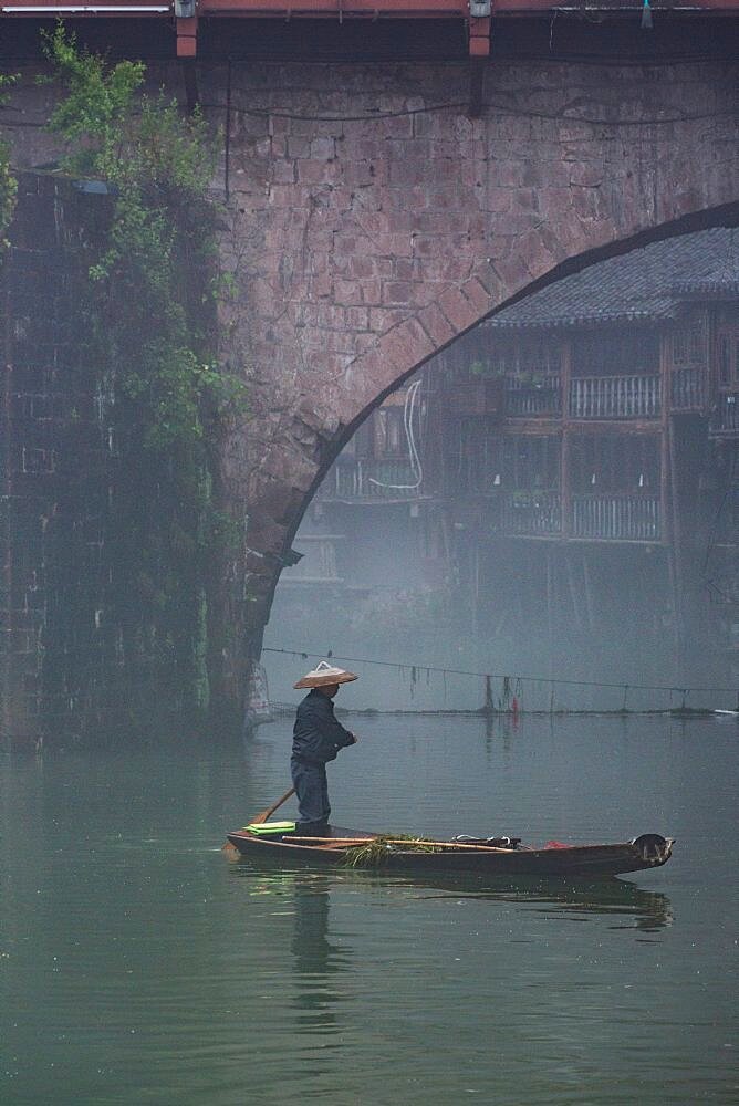 A man paddles a sampan in the fog by the Phoenix Hong Bridge on the Tuojiang River, Fenghuang, China.