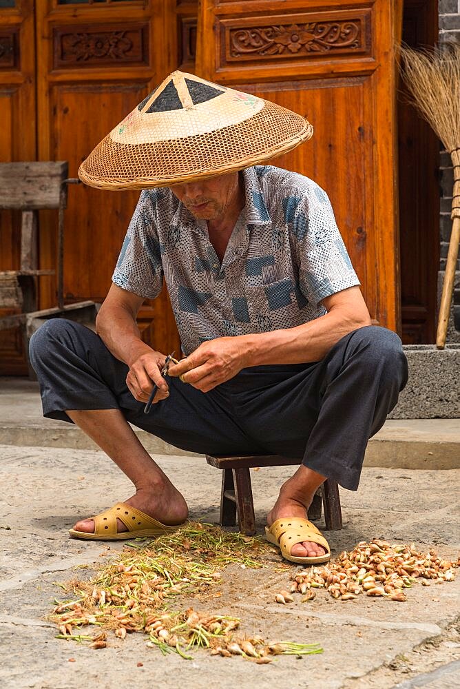 A Chinese man in a conical hat trims small onions for cooking in the ancient town of Furong, China.