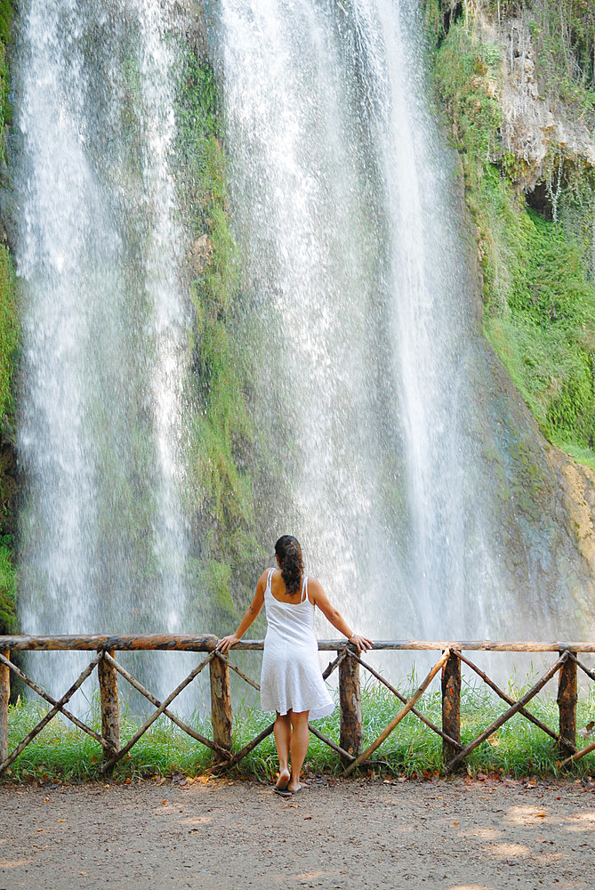 Young attractive brunette woman, wearing a white dress, relaxed and serene in a beautiful natural place, in front of a big waterfall, among assorted vegetation, Monasterio de Piedra, Zaragoza, Spain