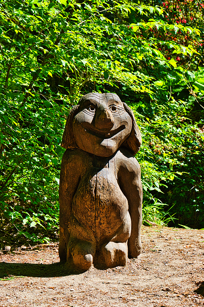 A wooden sculpture of a smiling cartoon-like character with a large head and short body, placed outdoors in a forested area.