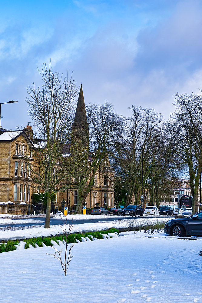 Snow-covered street scene featuring stone buildings, bare trees, parked cars, and a church spire. The sky is partly cloudy, light blue in Harrogate, UK.