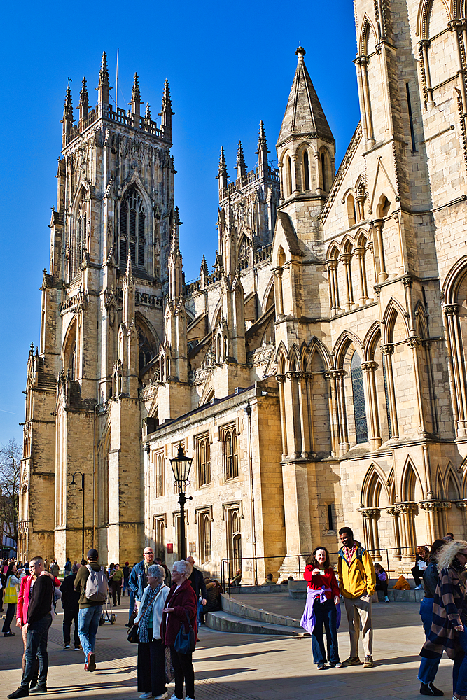 A view of York Minster with a crowd of people. The cathedral is made of light stone, set against a clear blue sky.