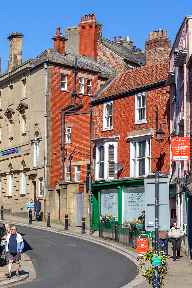 Street scene featuring brick buildings, people walking, and commercial storefronts under a bright, clear sky in Ripon, UK.
