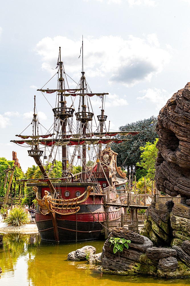 A large, decorative pirate ship is docked in a green pond with detailed rock formations and lush green foliage at Disneyland Paris.