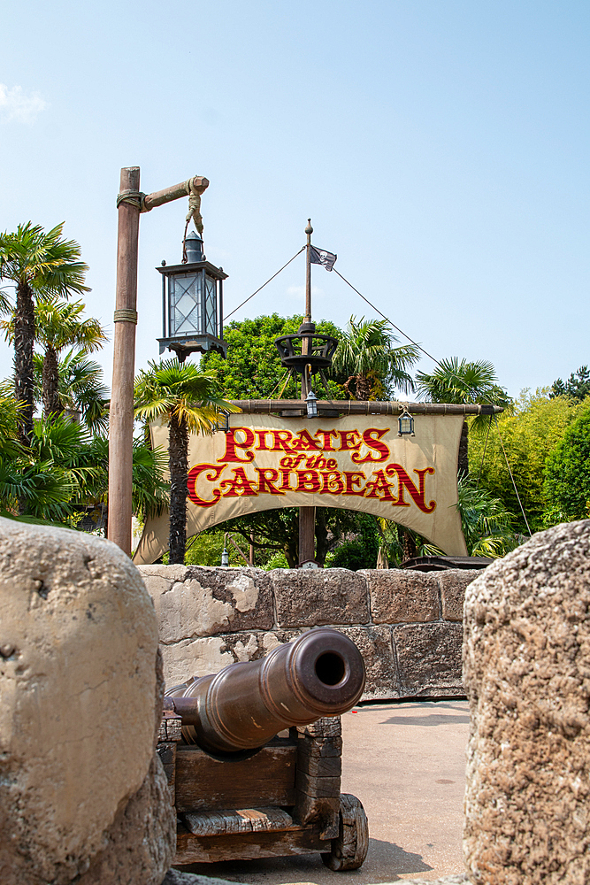 View of 'Pirates of the Caribbean' ride with cannon in foreground, sign in mid-ground, foliage, and blue sky at Disneyland Paris.