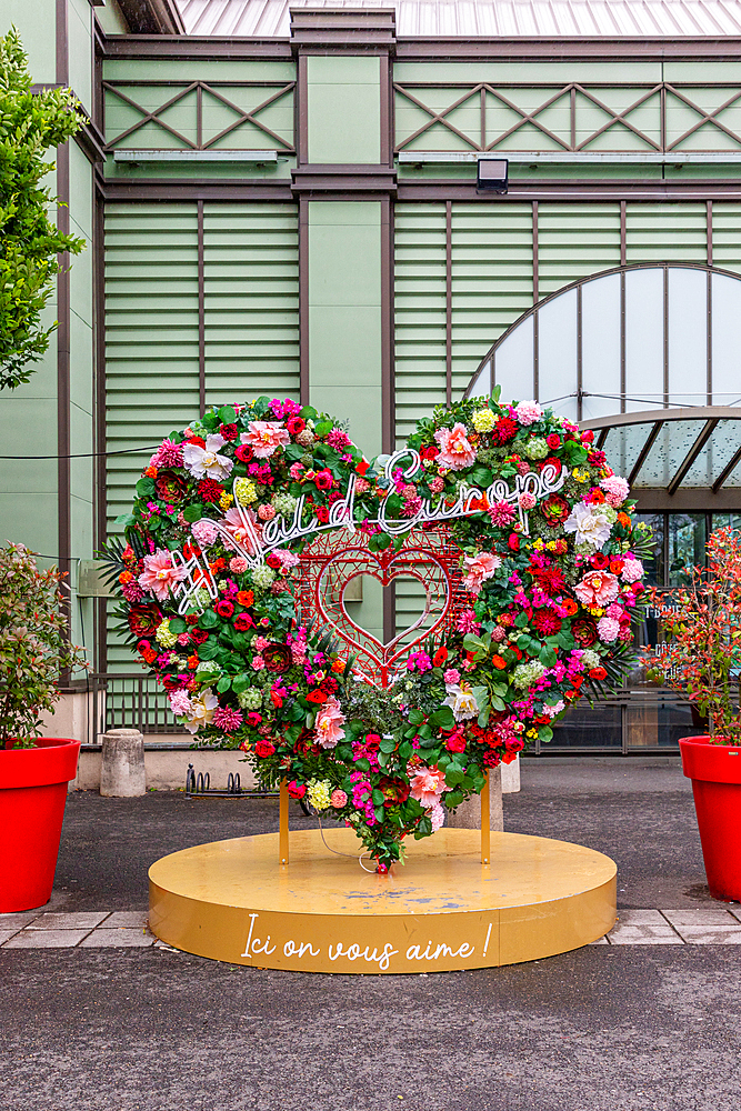 Floral heart sculpture with 'Val d'Europe' signage, standing on a gold platform with text, against a green and brown building.