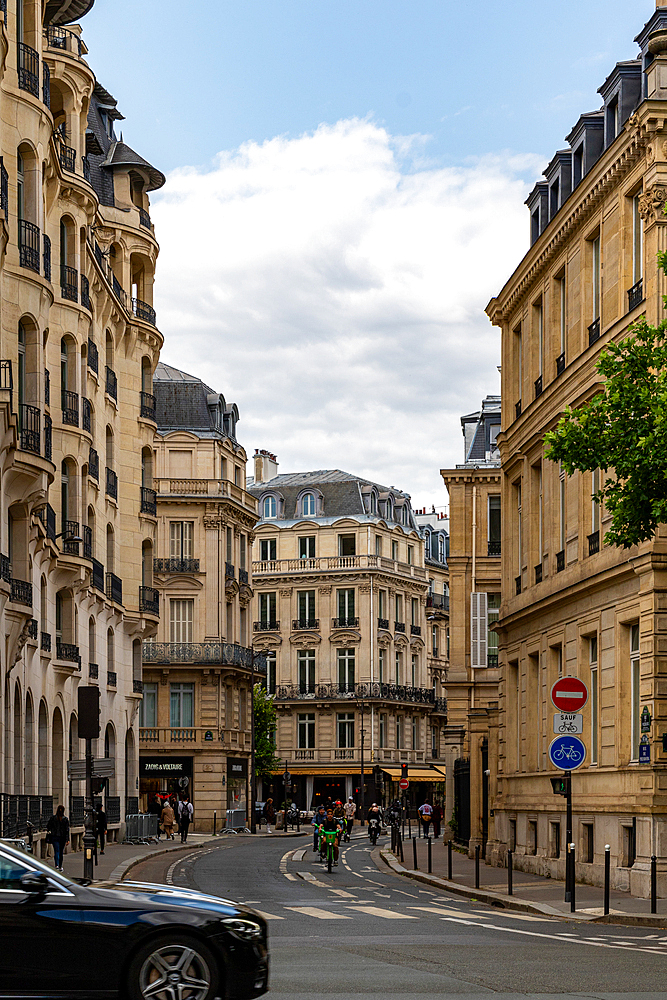 Street view of Parisian buildings lining a road with traffic and pedestrians, captured on a cloudy day.