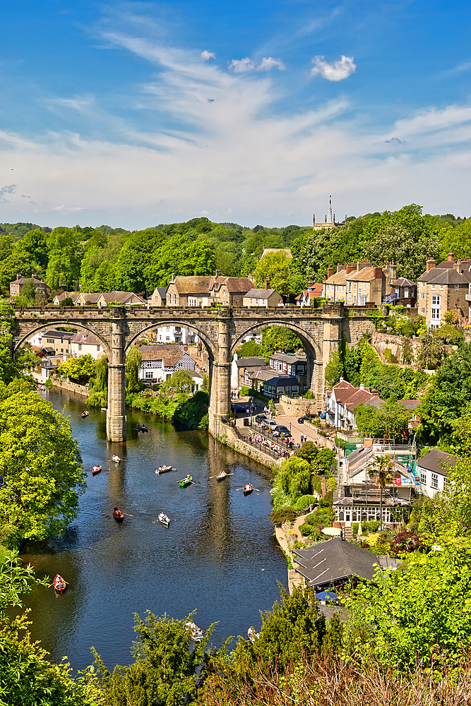 Scenic view of a stone viaduct spanning a river, with buildings, trees, and boats visible on a sunny day.