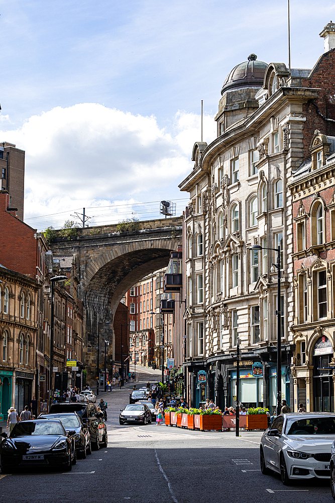 A sunlit street in a city, featuring a large stone railway arch, ornate buildings, and cars. The sky has scattered clouds in Newcastle upon Tyne, UK.