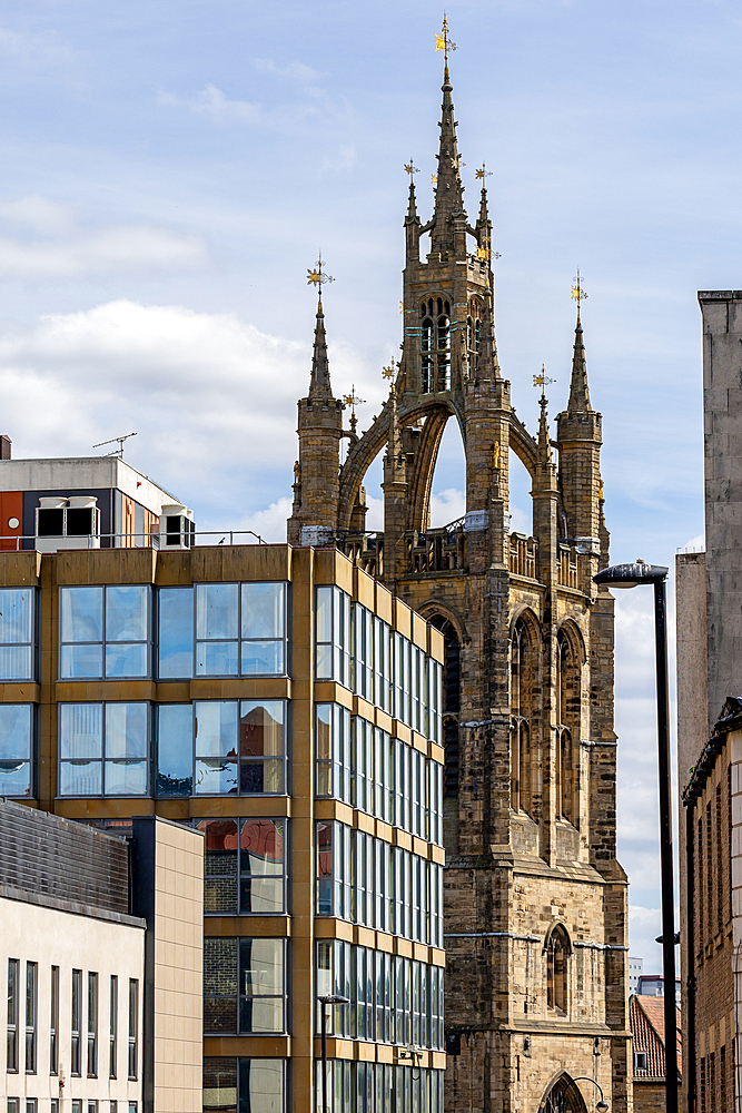 A Gothic tower with gold finials stands beside modern buildings featuring glass facades and concrete structures under a cloudy sky in Newcastle upon Tyne, UK.