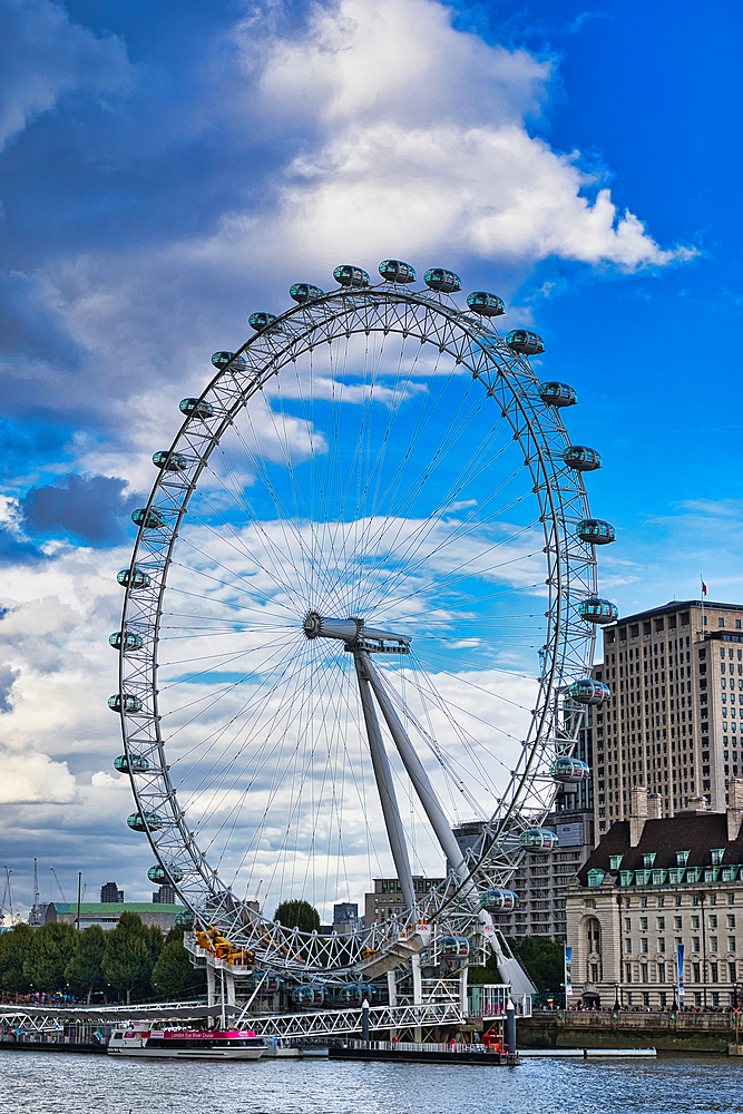 The London Eye dominates the frame, a colossal Ferris wheel against a dynamic sky of blue and grey clouds. Buildings and water are visible below.