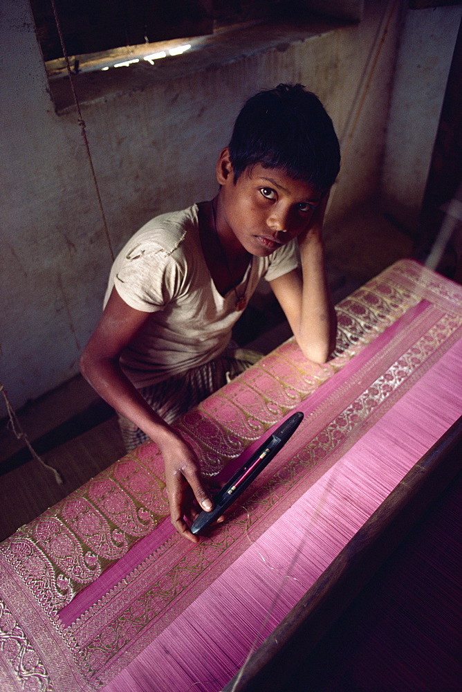 Young boy looking unhappy, sitting at loom working on silk material with gold thread, India, Asia