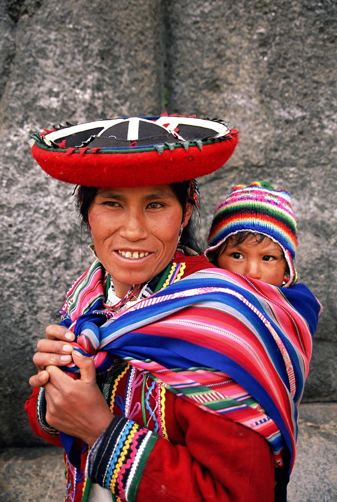 Portrait of a local woman in traditional dress, carrying her baby on her back, in front of Inca ruins, near Cuzco, Peru, South America